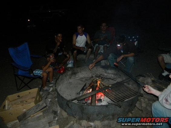Campfire scene at night with people sitting around a fire pit, some holding food