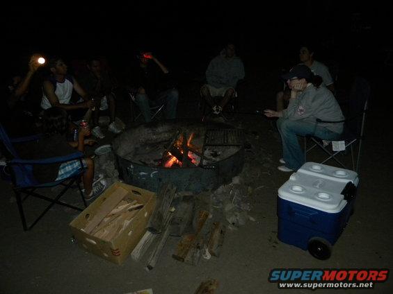 Group of people gathered around a campfire at night with a cooler nearby