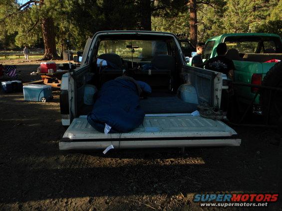 Pickup truck bed with a blue sleeping bag and camping gear. Another green truck is parked nearby