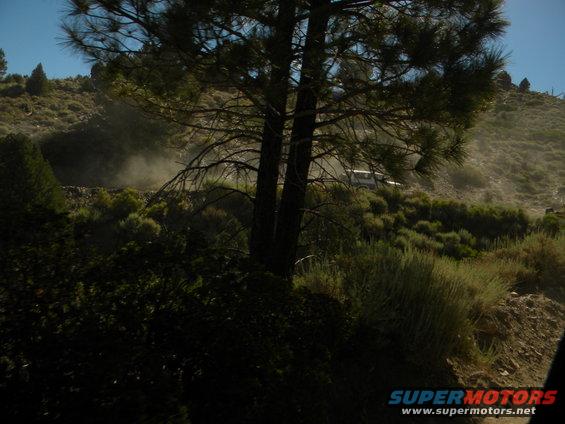 White SUV driving on a dusty, hilly dirt road with trees and brush