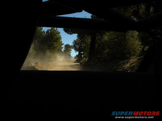 SUV driving on a dusty dirt road through a forest, seen from inside another vehicle