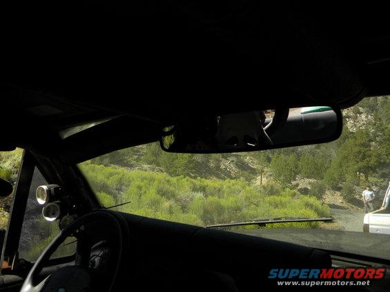 Interior view from a vehicle, looking out the windshield at a dirt road and green hills. Another vehicle is visible on the road