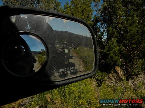 View in side mirror shows trucks on a dirt road with trees and hills