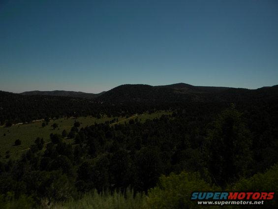 Wooded hills under a clear blue sky. A dirt road winds through the landscape