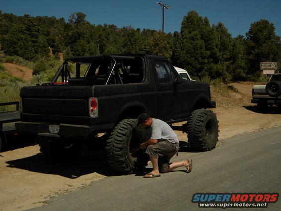 Man kneeling to change a large tire on a lifted black Ford Bronco. Another vehicle is parked behind it