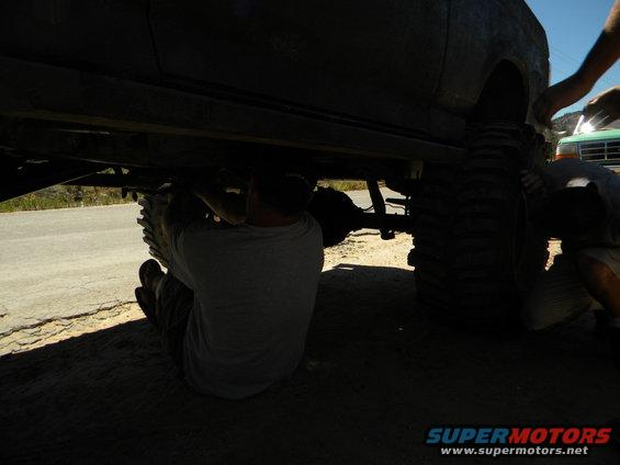 Two people working underneath a lifted 4x4 vehicle with large tires. Another vehicle is partially visible in the background