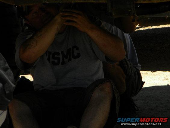 Two people are working underneath a vehicle. One person wears a grey t-shirt with 'USMC' on it