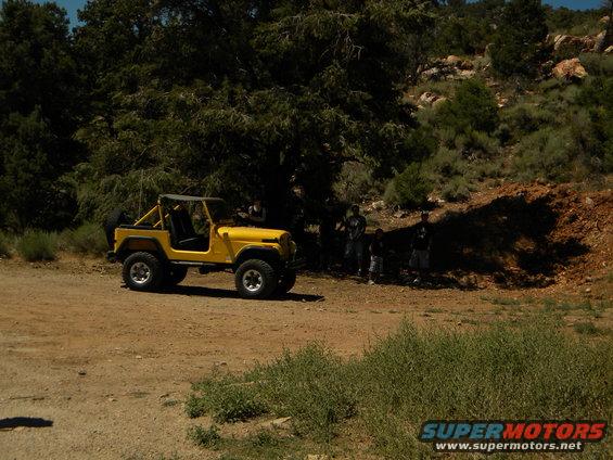 Yellow Jeep CJ with a roll cage and soft top parked on a dirt road with trees and hills in the background