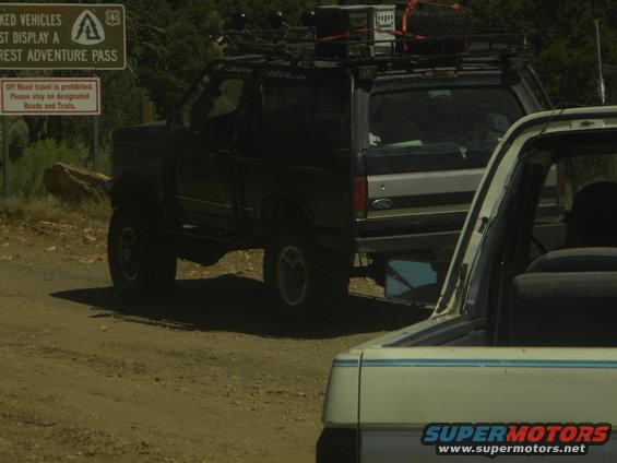1992 Ford Bronco with roof rack parked on a dirt road, another vehicle behind it. Signage visible