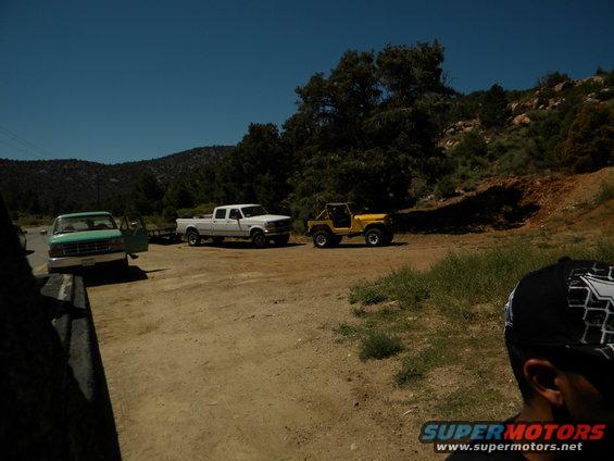 Green pickup, white pickup towing a trailer, and a yellow Jeep parked on a dirt road with trees and hills in the background