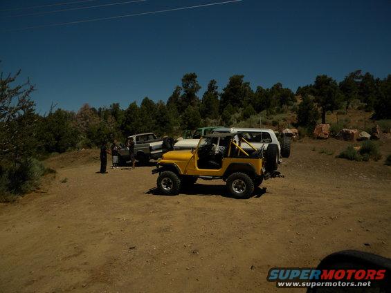 Yellow Jeep CJ-7 and other SUVs parked on dirt road with trees and hills in background