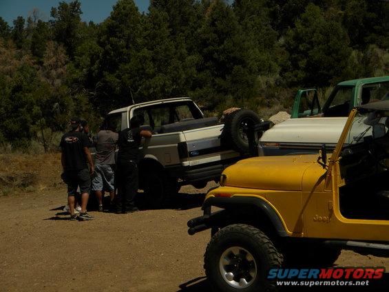 White Ford Bronco with spare tire on back, parked next to yellow Jeep CJ and green truck. People standing around