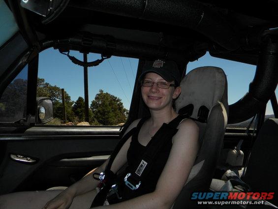 Woman wearing a cap and glasses sits in the passenger seat of an off-road vehicle, wearing a racing harness. The sky is blue with some trees visible outside