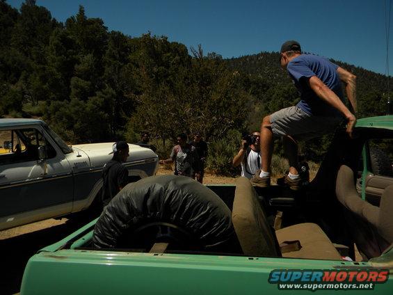 Green truck bed with spare tire, white and blue truck beside it, people in a wooded area