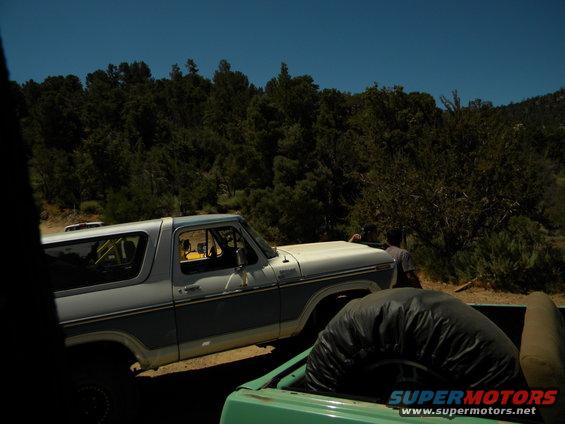 Side view of a light blue and white Ford Bronco parked next to a green pickup truck with a spare tire