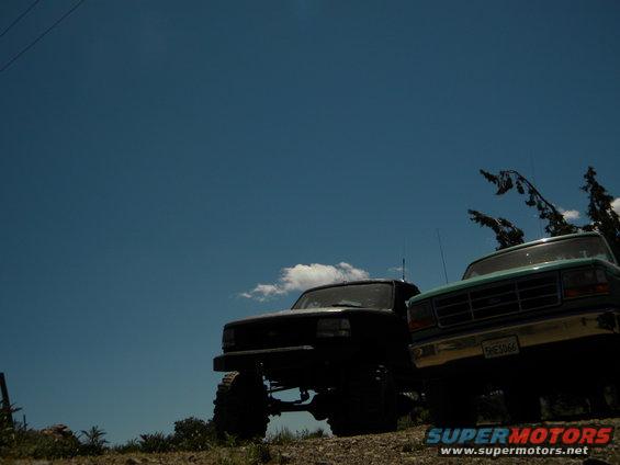 Black 1992 Ford Bronco and a teal 1990s Ford F-Series truck parked on a dirt incline under a blue sky