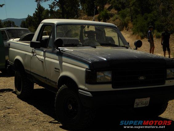 White and black Ford Bronco parked on a dirt road with another vehicle to its left. People stand in the background