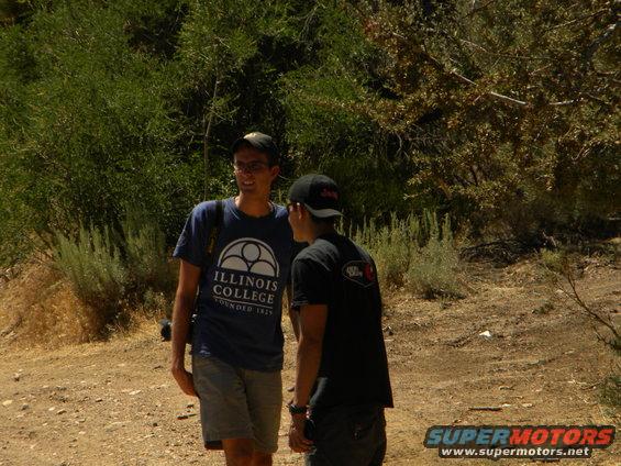 Two young men stand in a dry, wooded area. One wears a blue t-shirt with "Illinois College" text