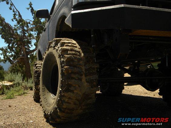 Off-road SUV with massive tires and a lifted suspension, parked on a dirt road with trees in the background