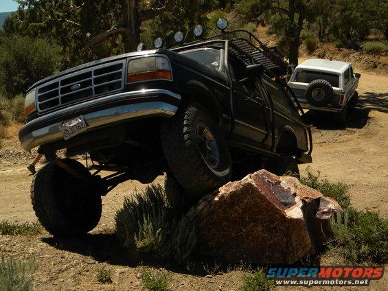 Black Ford truck with lifted suspension driving over a rock, another white SUV in background