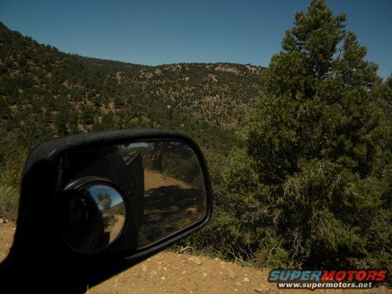 Side mirror reflection shows a dirt road winding through a wooded, hilly landscape under a clear blue sky