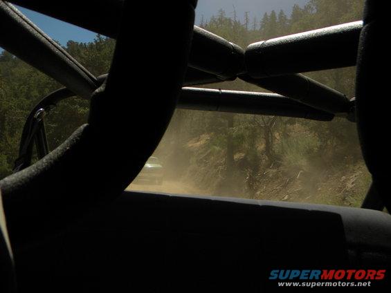 View from inside a vehicle looking out at another vehicle kicking up dust on a dirt road