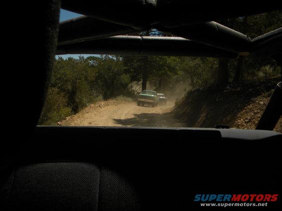 Two trucks drive on a dusty dirt road through trees. One truck is green