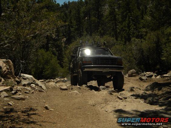1992 Ford Bronco driving over rocks on a dirt path with trees in the background
