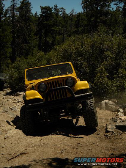 Yellow Jeep CJ off-roading on a rocky trail with trees in the background