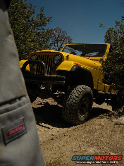 Yellow Jeep on a dirt path, front right tire visible, brush on the right