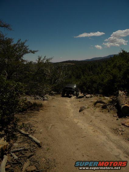Dark 1992 Ford Bronco on a dirt road, angled slightly from the front, with trees and hills in the background