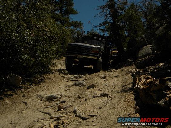 Truck climbing a rocky trail in the woods. Another vehicle is visible behind it
