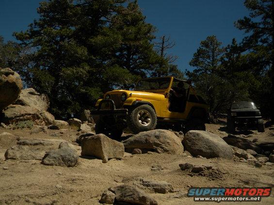 Yellow Jeep Wrangler on rocky terrain, driver visible, black SUV in background