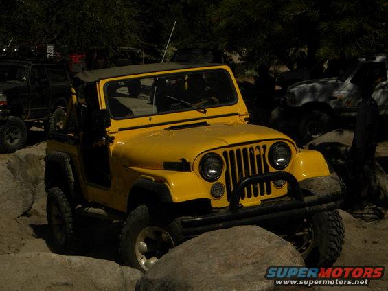 Yellow Jeep CJ driving over rocks, with a black truck and a white truck in the background