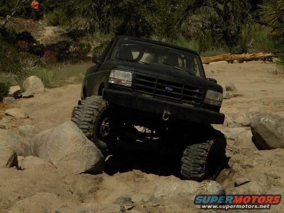 Black SUV crawling over rocks on a dirt trail. Someone is standing on a hill in the background