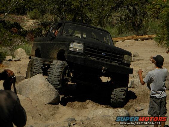 Black truck with big tires going over rocks. Guy in hat watching