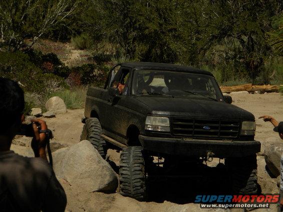 Black 1992 Ford Bronco with large tires on a rocky trail, driver visible, front-right view