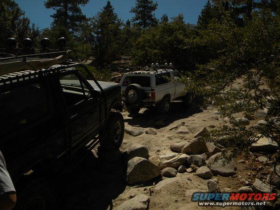 Dark off-road SUV in foreground, white SUV on rocky trail behind it