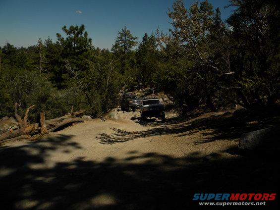 Two dark SUVs on a rocky dirt road in a wooded area, one in the foreground, one behind