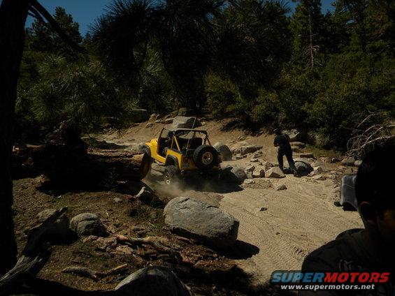 Yellow Jeep Wrangler driving over rocks on a dirt trail, with a person walking nearby