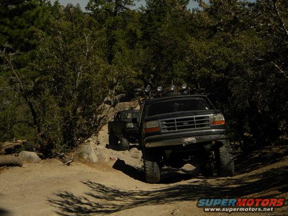 Black 1992 Ford Bronco with off-road lights on roof rack, driving on a dirt trail with rocks and trees