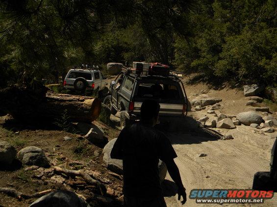 Two SUVs driving on a rocky dirt trail in a forest. One is dark colored, the other is silver