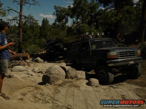 Dark 1992 Ford Bronco on a rocky trail, front passenger side visible, with another dark SUV behind it