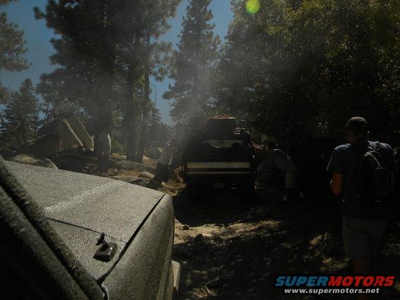Textured hood of a dark SUV in the foreground, with a 1992 Ford Bronco and other vehicles on a rocky, wooded trail