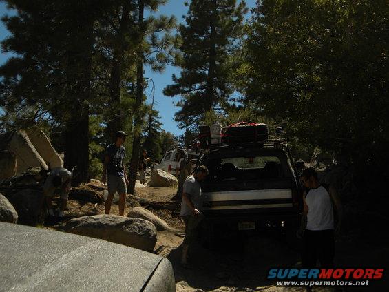 Off-road vehicles on a rocky trail with people around. One vehicle has a roof rack with gear