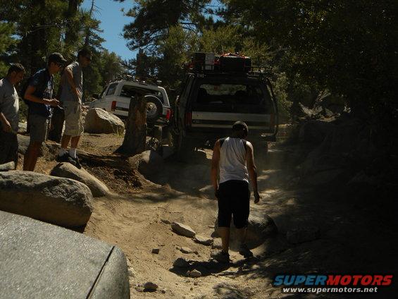 1992 Ford Bronco, dark color, rear view, on a rocky trail with roof rack