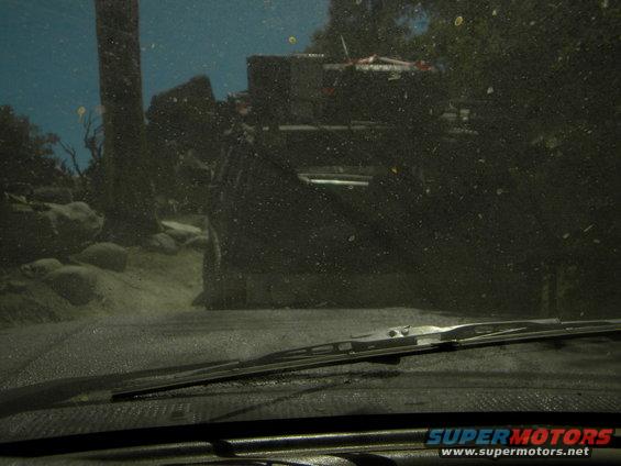 View from inside a vehicle looking out at another vehicle on a dirt road with trees and rocks