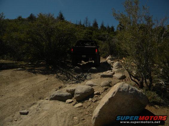 Black pickup truck driving over rocks on a dirt trail with trees and bushes around