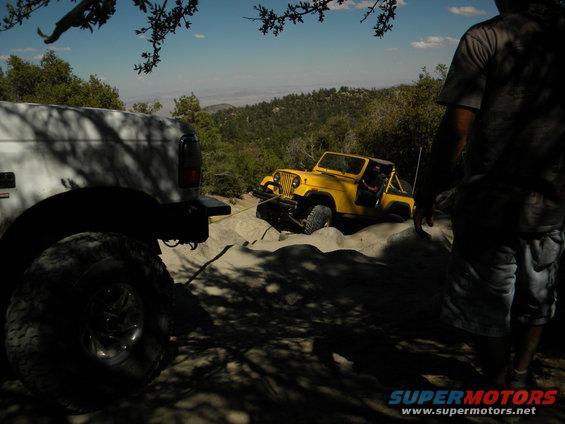 White vehicle in foreground, yellow Jeep stuck on rocks with tow rope attached. Person standing on right