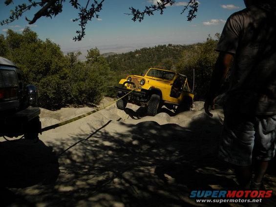 Yellow Jeep being pulled by a tow strap attached to a dark SUV, on a sandy, off-road trail